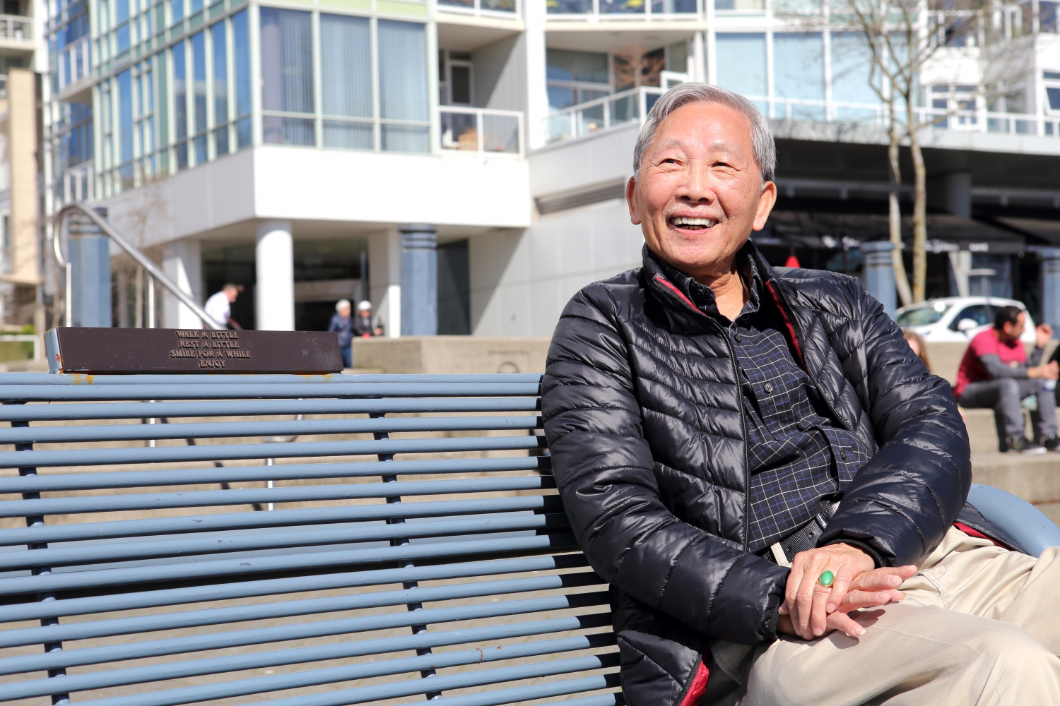 Smiling Asian man in a puffy jacket sitting on a grey-blue bench in the sun