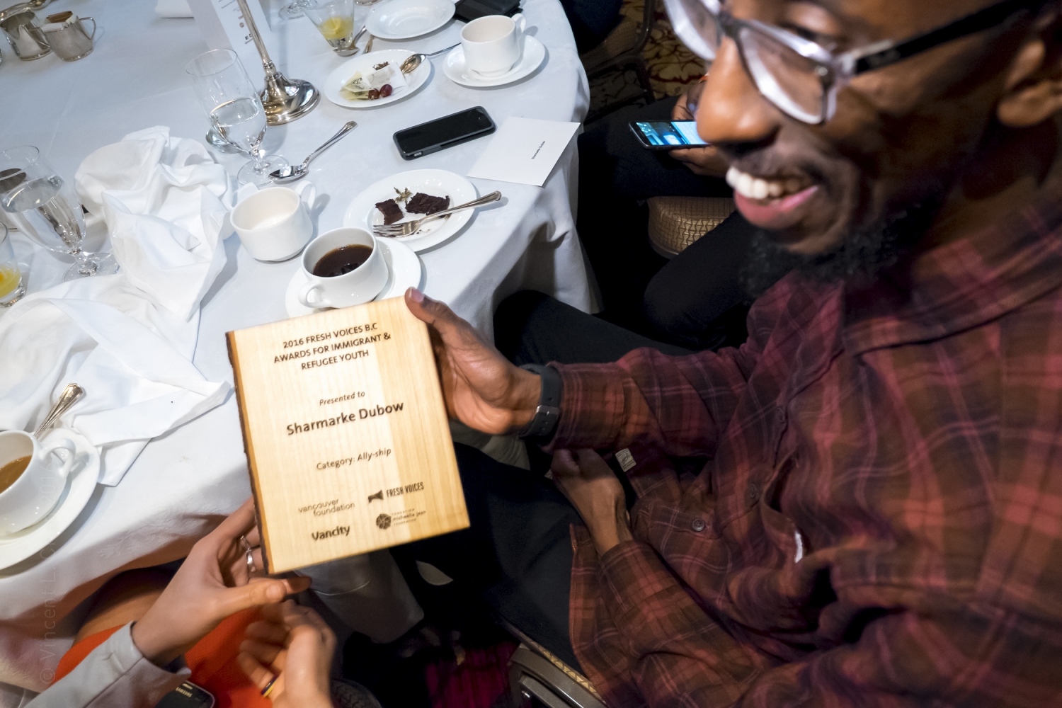 Smiling man sitting at a banquet table holding a wooden award plaque with words "2016 Fresh Voices B.C. Awards for Immigrant and Refugee Youth presented to Sharmarke Dubow"