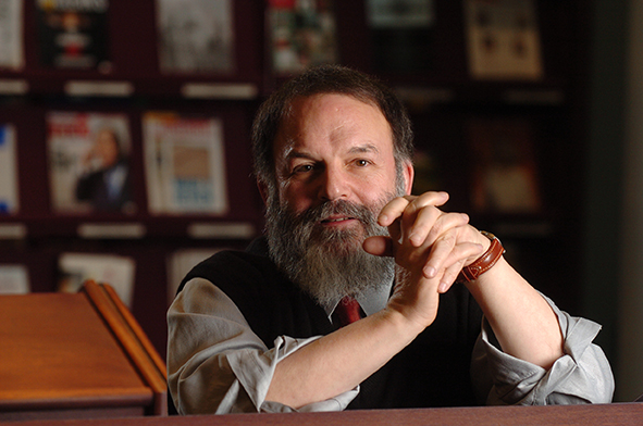 An older white man with grey hair and beard wearing a blue shirt and dark vest, sitting at a wooden table, hands clasped together