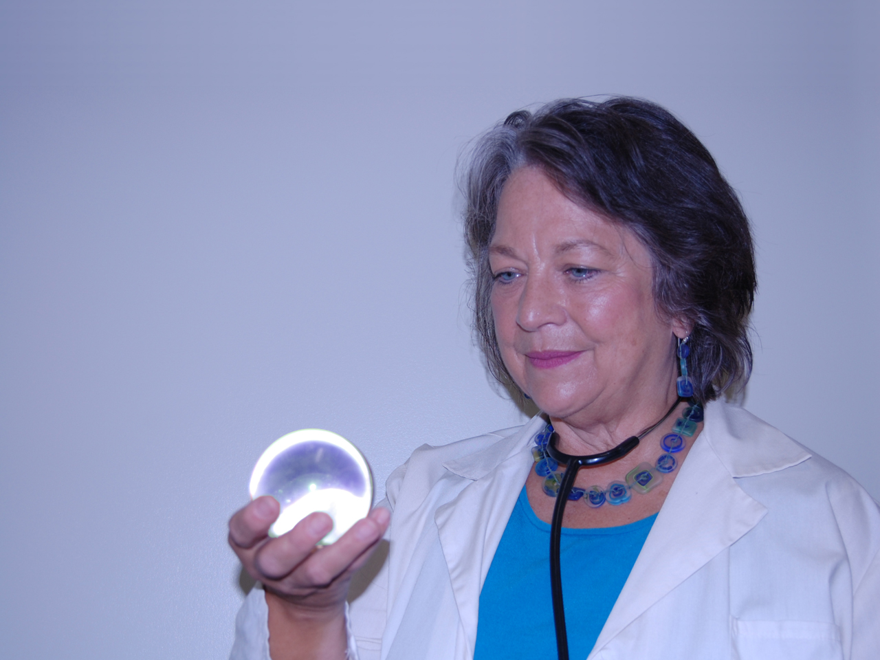 A white woman with grey hair and blue eyes wearing blue jewelry, a turquoise shirt and white lab coat, holding a glass orb with a stethescope around her neck