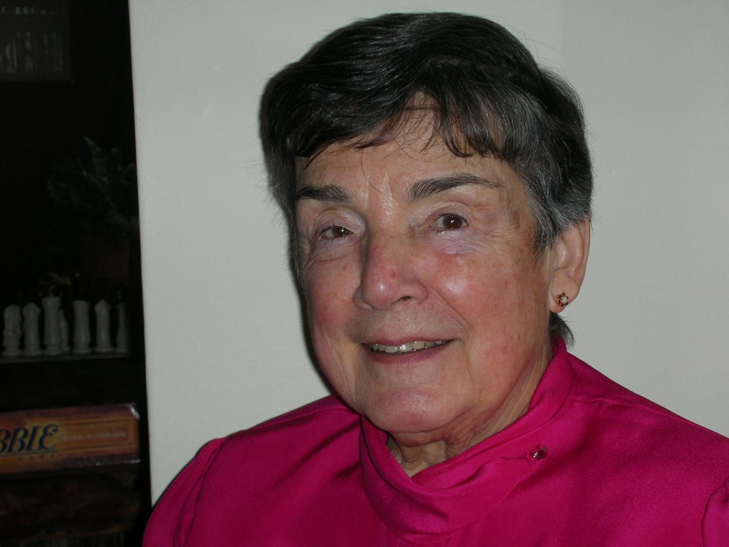 An elderly white woman with dark grey hair and brown eyes, wearing a magenta top and stud earrings, smiling