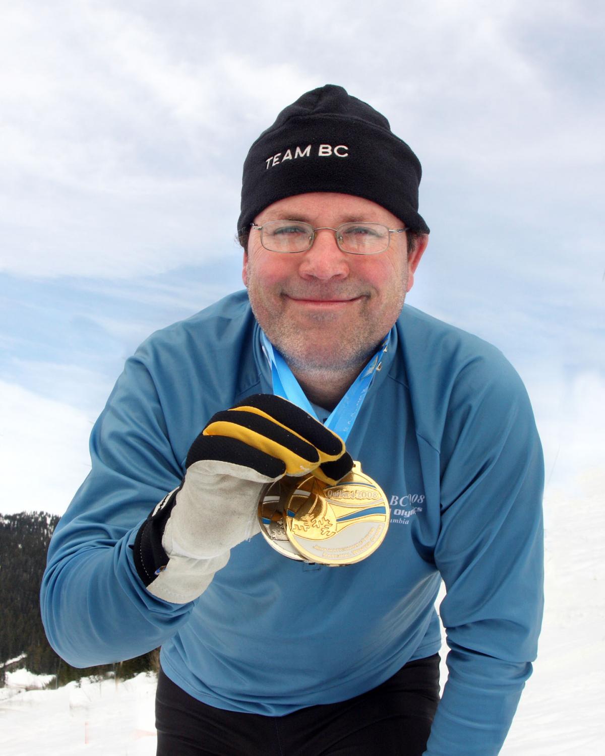 A smiling middle aged white man with glasses and light facial stubble wearing a jacket and black toque holding two Special Olympics medals