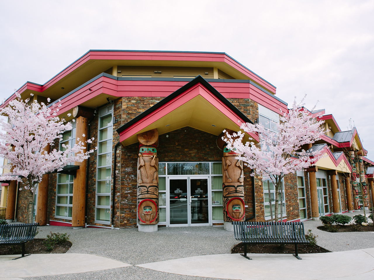 Outside view of a brick building with red-accented roofing, double glass doors flanked by two carved and painted indigenous totem-pole woodworks