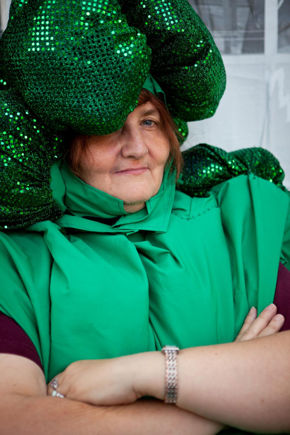 A woman in a green costume with a dark green, sequined bulbous hat resembling broccoli florets, smiling slightly with crossed arms