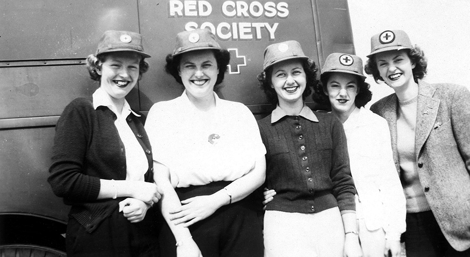 A 1940s era black and white photo of a smiling group of young women in front of a van with the words "Red Cross Society" on it