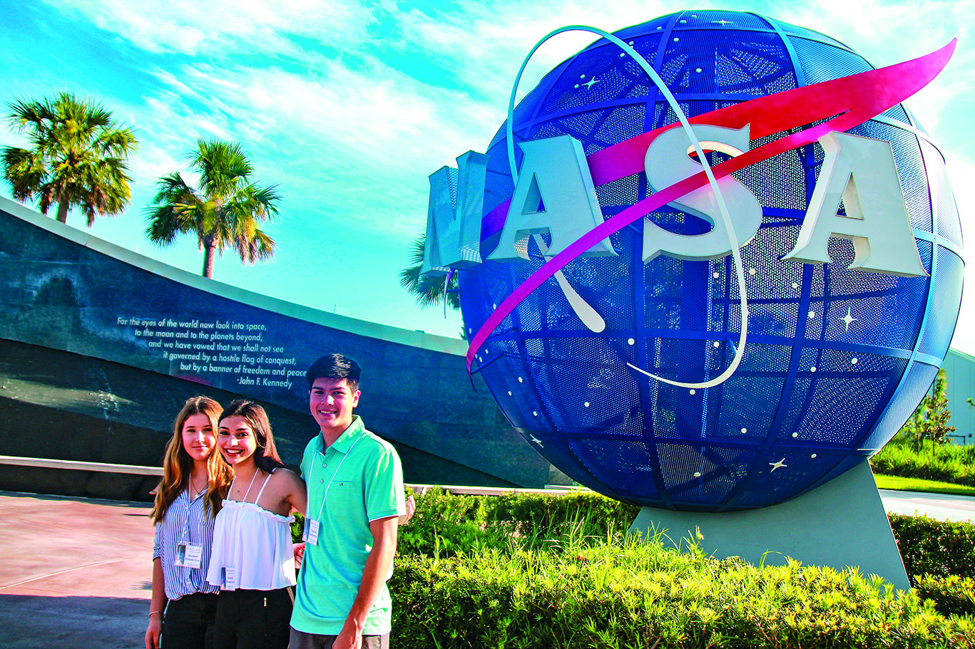 Three smiling students with badges standing in front of an sculpture of the NASA logo: a blue sphere with NASA in white blocky font and a red stylized arrow