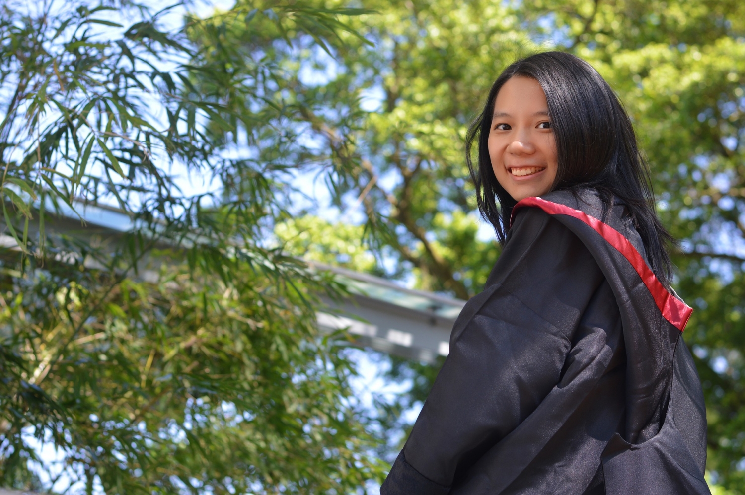 Young asian woman in a black graduation gown with red trim, smiling with green leafy foliage in the background
