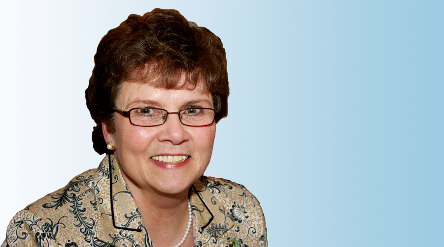 An older white woman with short red-brown hair and glasses, wearing a floral patterned blouse and smiling, against a neutral, blue-toned gradiant background