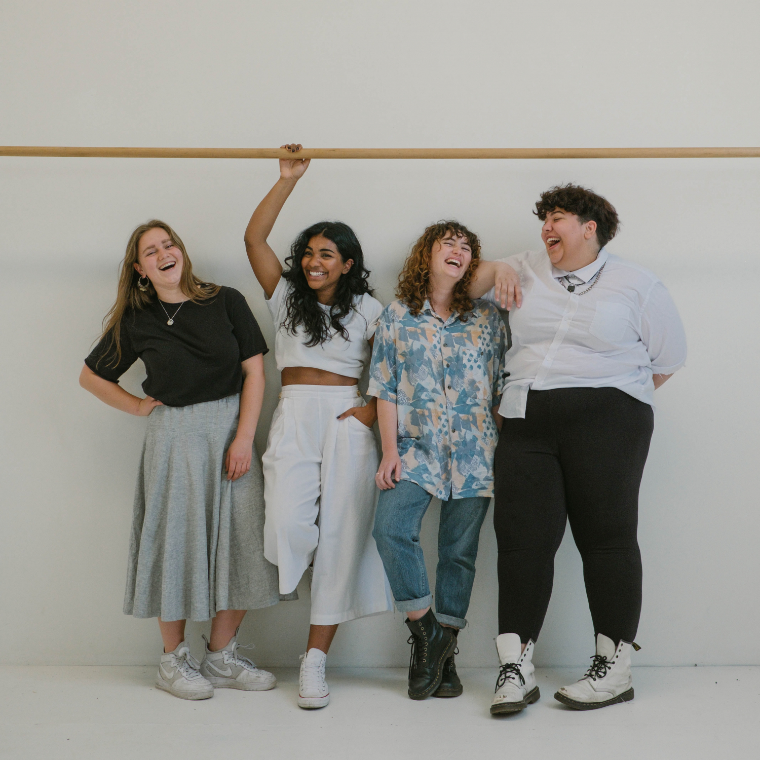 Four laughing friends in casual wear leaning against a white wall with a bar of wood running horizontally overhead. Credit Emma Chau Tran from Unsplash
