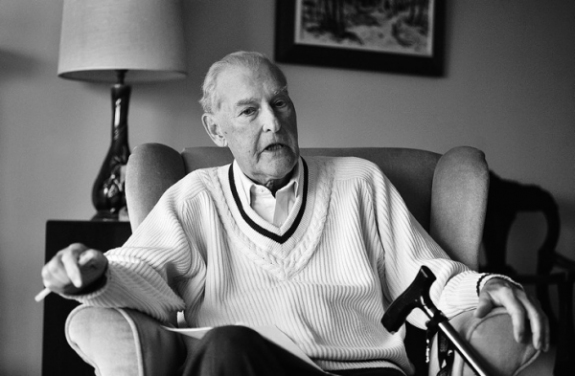 A black and white picture of an elderly man with a cane, wearing a v-neck braided collar sweater, sitting in a wingbacked chair