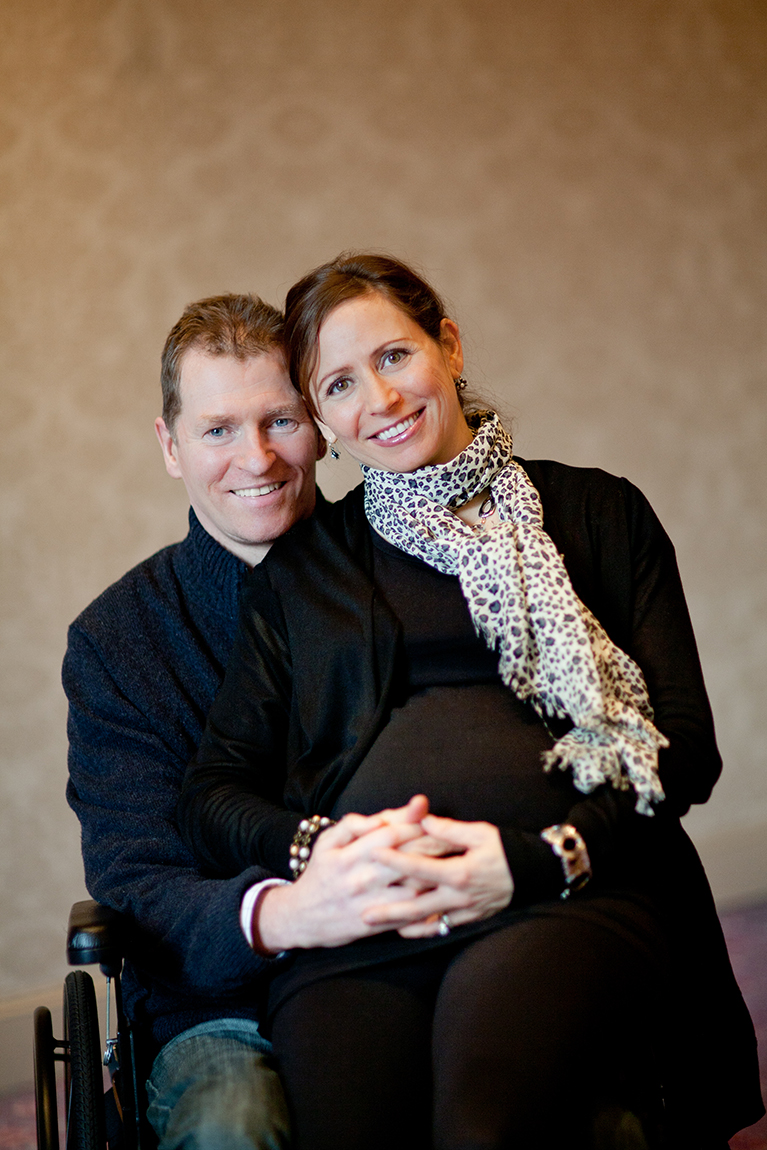 A smiling brown haired woman sitting in the lap of a smiling man in a wheelchair