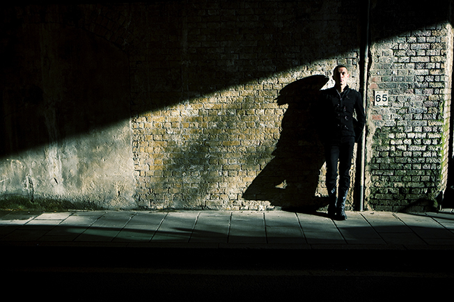 A man in dark clothing leaning against a brick wall in low light, with his shadow cast next to him